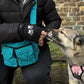 Das Model präsentiert Gassi-Tasche, Bauchtasche sowie Umhängetasche aus Canvas-Stoff, türkis, mit vielen kleinen hellblauen Pfoten. Neben ihr sitzt ein Hund, der aus ihrer Hand ein Leckerli frisst. An den Händen trägt das Model Nijens Handstulpen aus Schurwolle schwarz mit zwei Pfotenstickerei.