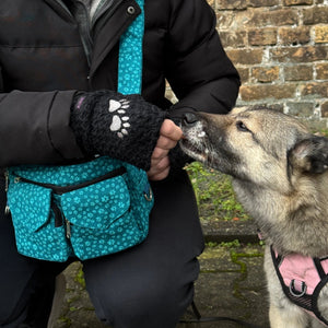 Das Model präsentiert Gassi-Tasche, Bauchtasche sowie Umhängetasche aus Canvas-Stoff, türkis, mit vielen kleinen hellblauen Pfoten. Neben ihr sitzt ein Hund, der aus ihrer Hand ein Leckerli frisst. An den Händen trägt das Model Nijens Handstulpen aus Schurwolle schwarz mit zwei Pfotenstickerei.