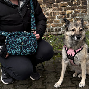 Ein Model präsentiert die Gassi-Tasche, Bauchtasche sowie Umhängetasche aus Canvas-Stoff, Dunkelblau mit vielen kleinen türkisenen Pfoten. Neben ihr sitzt ein Hund. An den Händen trägt das Model Nijens Handstulpen aus Schurwolle schwarz mit zwei Pfotenstickerei.