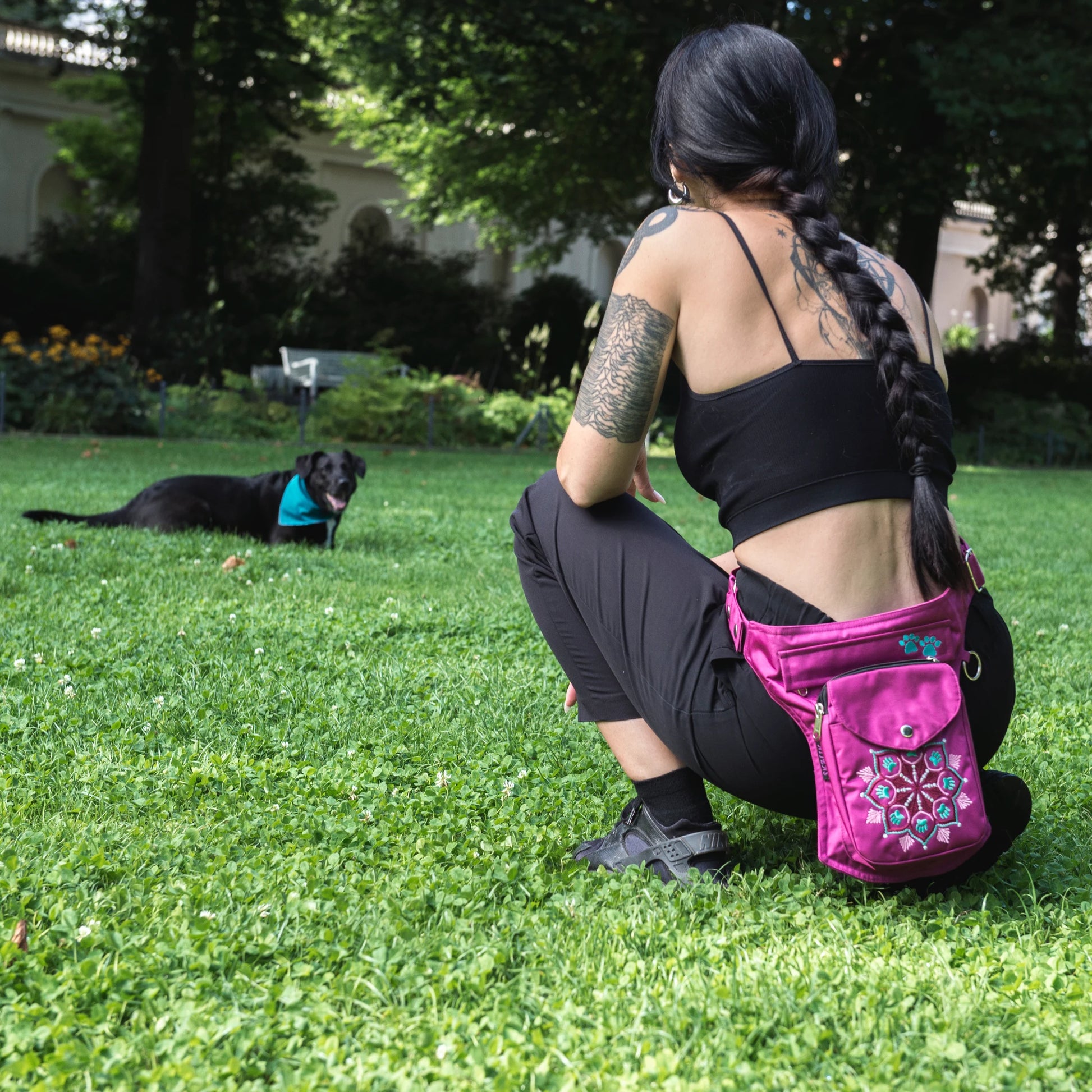 Gassi-Tasche, Bauchtasche und Gürteltasche aus wasserabweisendem Nylonstoff in Pink mit Pfoten-Mandalastickerei. Diese Gürteltasche hat zwei Reißverschlussfächer vorne sowie ein Einsteckfach, welches durch einen Überschlag mit Druckknopf verschlossen wird. Auf der hinteren Seite befindet sich ein weiteres Reißverschlussfach. An der Seite des Taschengurtes befindet sich ein kleiner Metallring zum Anhängen kleiner Beuteltaschen. Eine Frau mit Hund.