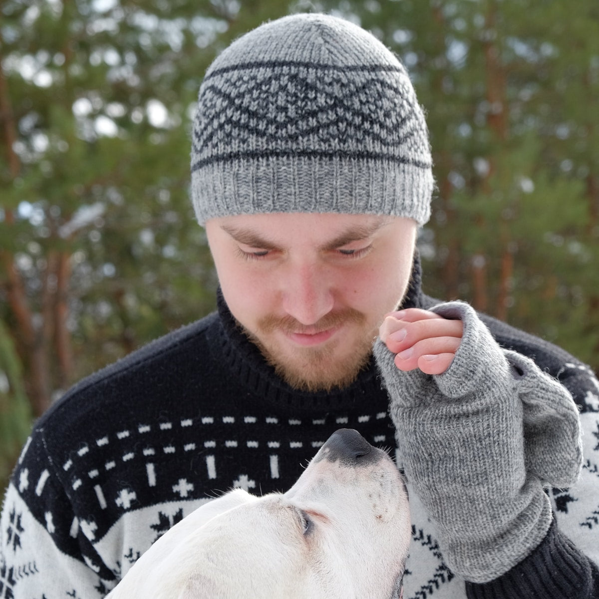 Mann trägt graue Strickmütze aus Schurwolle mit schwarzem Muster und passende Pulswärmer beim Spaziergang mit seinem Hund im Wald.