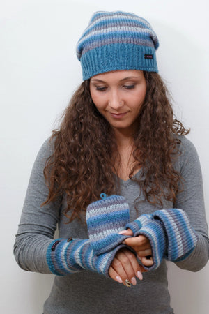 Das Model präsentiert eine gestreifte long Beanie Mütze aus Schurwolle in Grau, Blautönen. An den Händen trägt sie fingerfreie Handschuhe aus Wolle gestreift in Blautönen, welche sich mit einer Fingerkappe zu Fäustlingen umwandeln lassen. Zwei Tragemöglichkeiten.