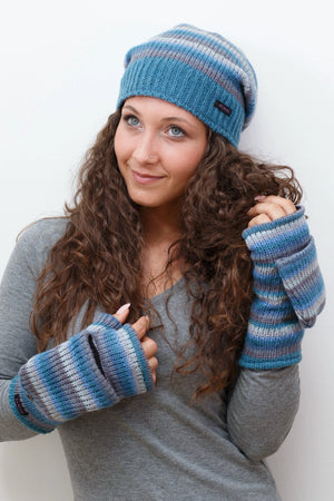 Model präsentiert eine gestreifte long Beanie Mütze aus Schurwolle in Grau, Blautönen. An den Händen trägt sie fingerfreie Handschuhe aus Wolle gestreift in Blautönen, welche sich mit einer Fingerkappe zu Fäustlingen umwandeln lassen. 