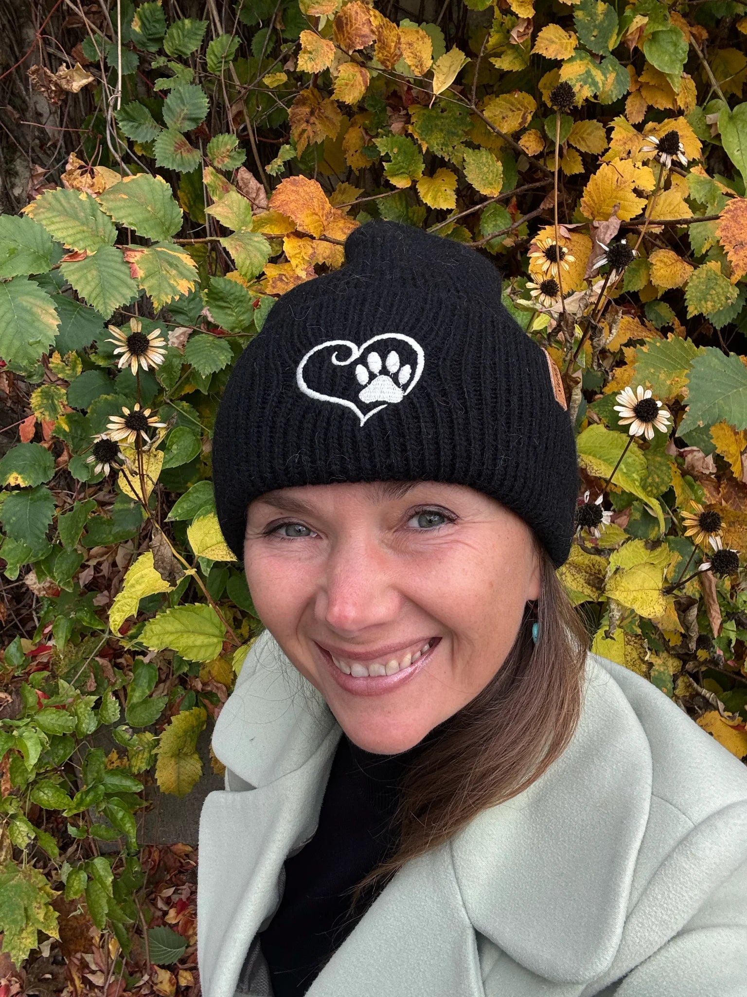 Nahaufnahme einer Frau mit schwarzer Nijens Angora Beanie Marseille und Pfote-&-Herz-Stickerei vor bunten Herbstblättern
