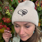 Nahaufnahme einer Frau mit Nijens Angora Beanie in Beige und Pfote-&-Herz-Stickerei vor bunten Herbstblättern