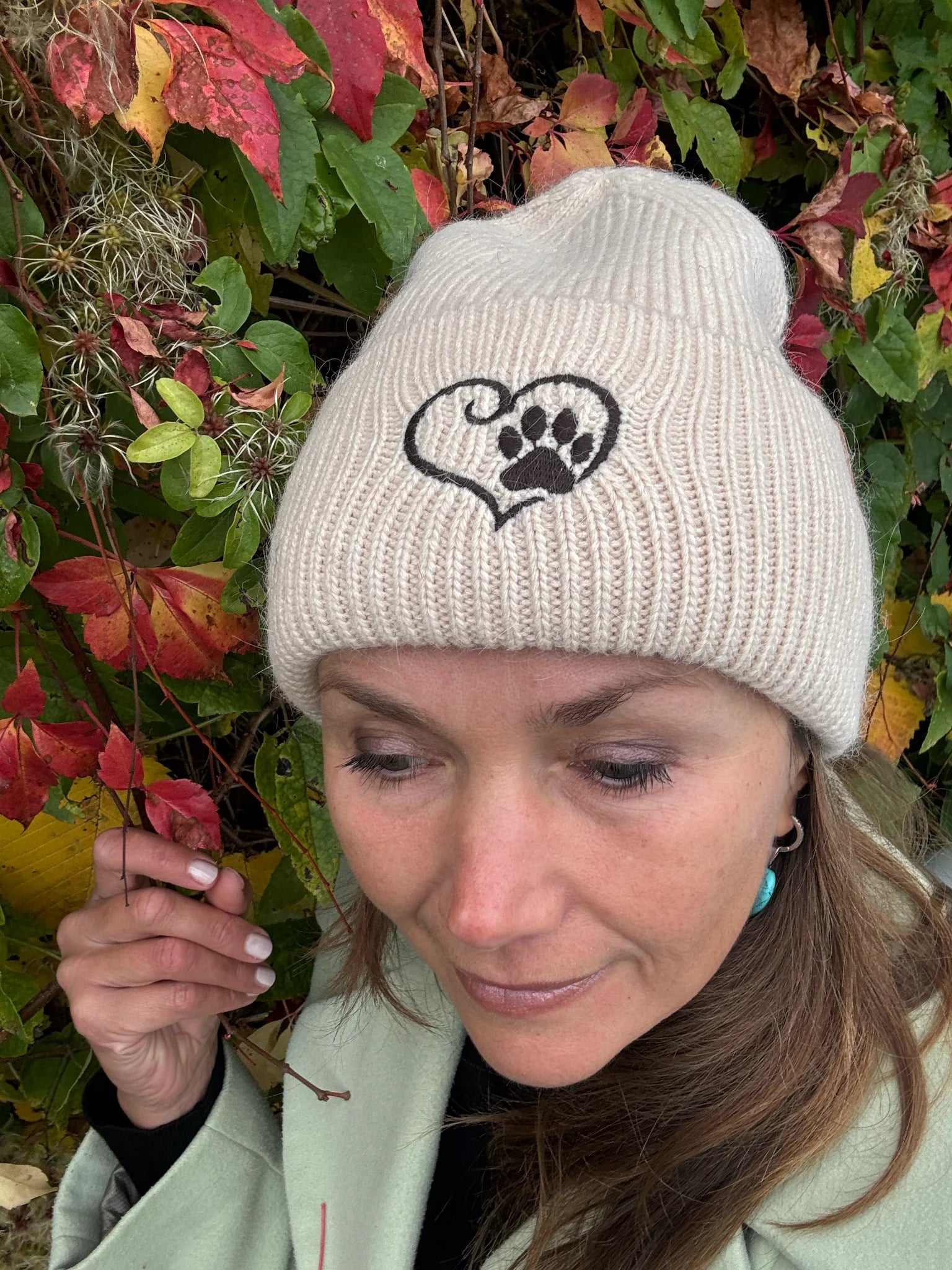 Nahaufnahme einer Frau mit Nijens Angora Beanie in Beige und Pfote-&-Herz-Stickerei vor bunten Herbstblättern