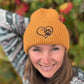Nahaufnahme einer Frau mit oranger Nijens Angora Beanie Marseille und Pfote-&-Herz-Stickerei vor bunten Herbstblättern