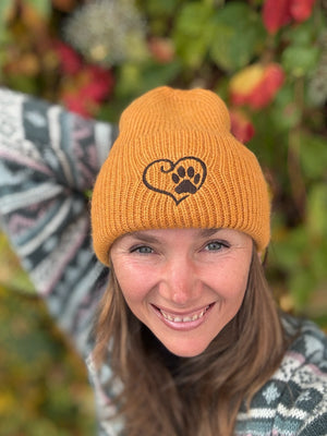 Nahaufnahme einer Frau mit oranger Nijens Angora Beanie Marseille und Pfote-&-Herz-Stickerei vor bunten Herbstblättern
