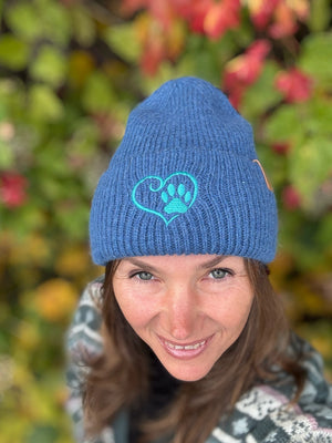 Nahaufnahme einer Frau mit blauer Nijens Angora Beanie Marseille und Pfote-&-Herz-Stickerei vor bunten Herbstblättern